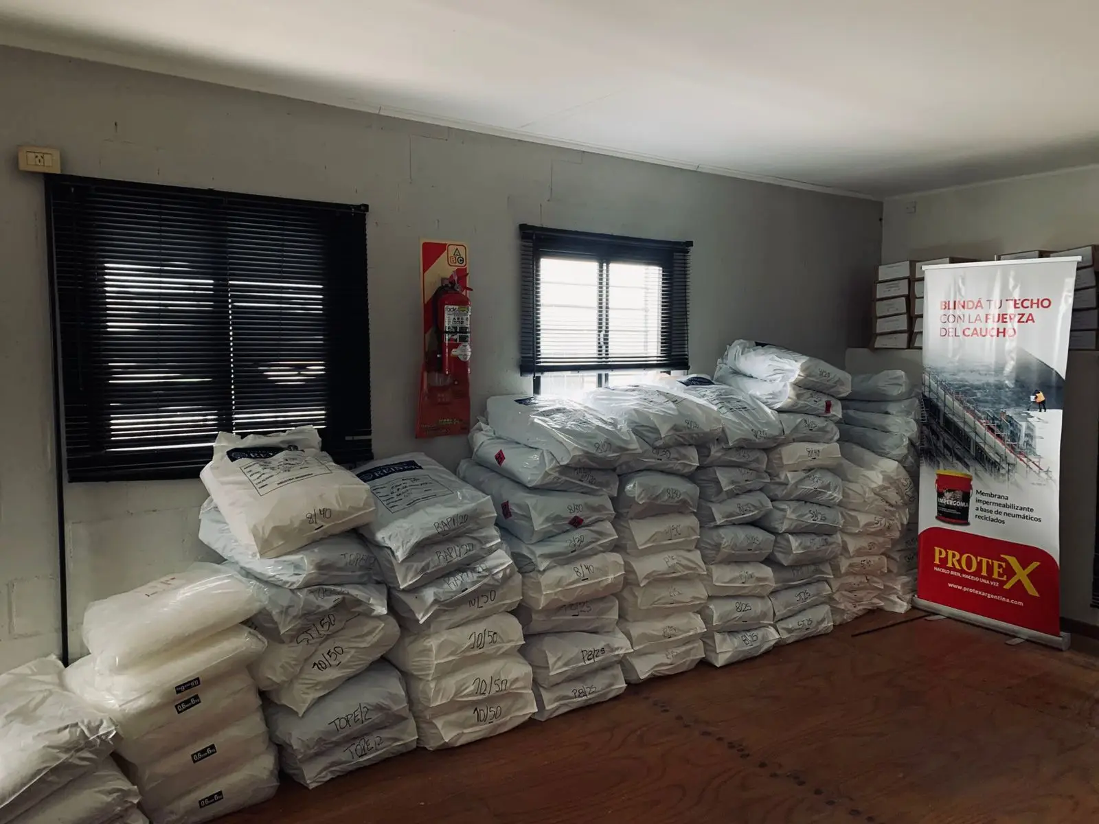 Depósito interno con bolsas de material apiladas y rotuladas a mano según su medida, junto a un banner de la línea de impermeabilizantes Protex.
