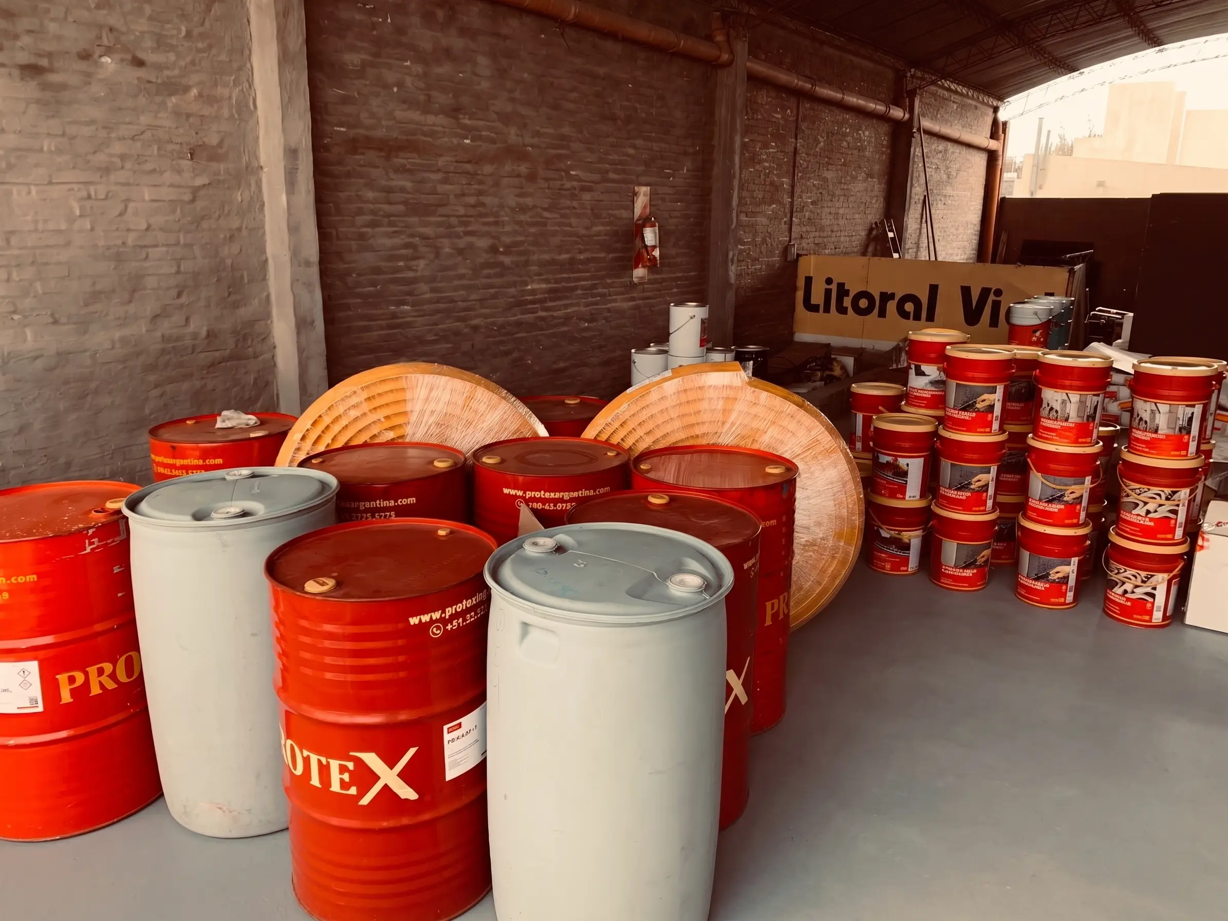 Sector de depósito con tambores de doscientos litros, rollos de membrana y baldes de productos Protex apilados sobre el piso de hormigón.